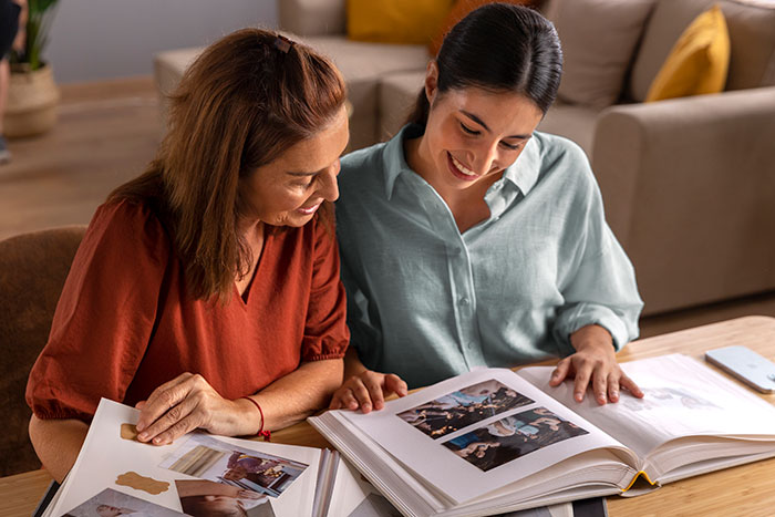 Two women smiling and looking through photo albums, discussing family moments and relationships at a table. Two women smiling and looking through photo albums, discussing family moments and relationships at a table.