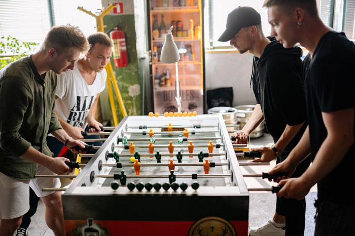 Group of young men playing foosball indoors, highlighting a woman telling drunk boyfriend to stop acting like a jerk.
