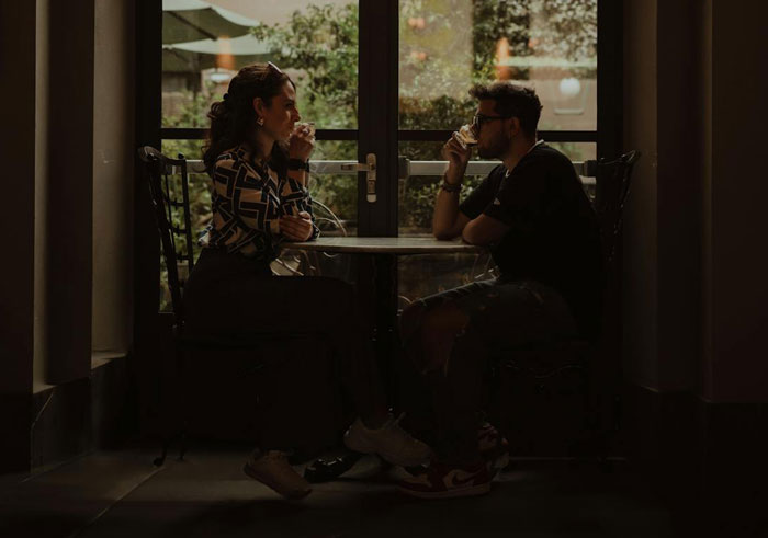 Woman and drunk boyfriend sitting at a table indoors, she looks upset as he drinks, tense relationship moment.