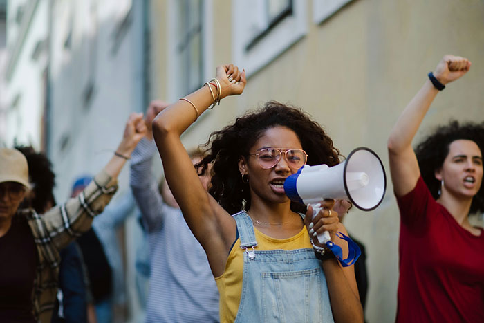 Woman wearing glasses and denim overalls uses megaphone leading a protest against beauty standards with raised fists. Woman wearing glasses and denim overalls uses megaphone leading a protest against beauty standards with raised fists.