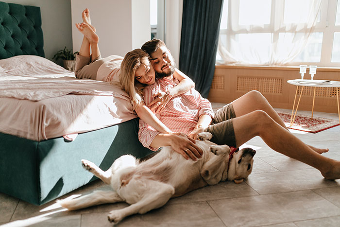 Couple playing with dog in sunlit bedroom, woman showing affection while man relaxes on the floor near bed.