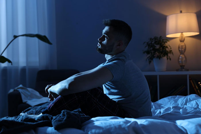 Man sitting on bed at night looking thoughtful, reflecting on woman party relationship partner issues in dim room.