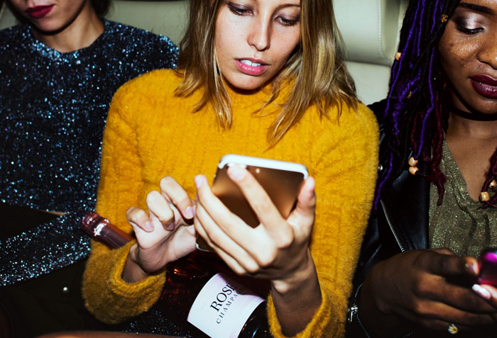 Woman at a party holding a champagne bottle and using a smartphone, surrounded by friends and potential relationship partners.