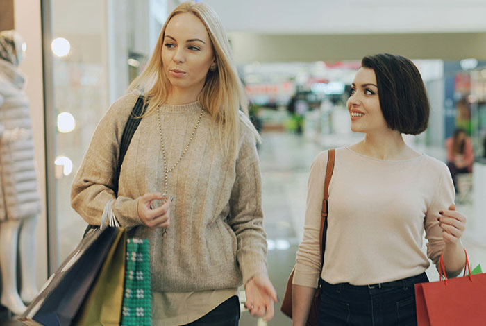 Two women shopping in a mall, one looking annoyed while the other smiles, representing attention-seeking mom behavior.