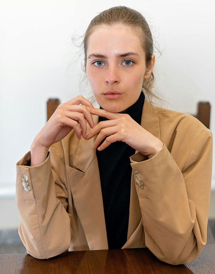 Young woman in a tan blazer and black shirt sitting at a table, reflecting on refusing to wear dresses for a wedding.