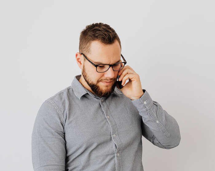 Man wearing glasses and a gray shirt looking concerned while talking on the phone about girlfriend refusing dresses for wedding.