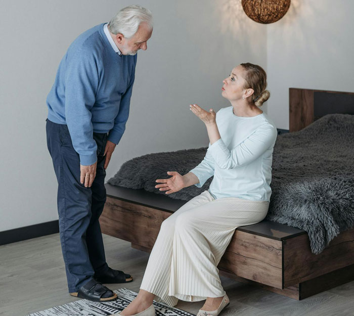 Woman sitting on bed speaking emotionally to an older man in a blue sweater about family secrets unfolding
