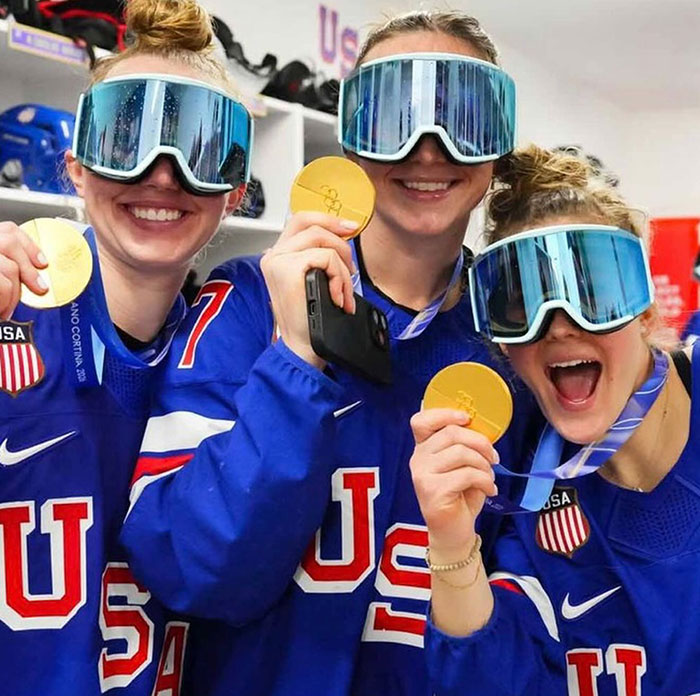 US women's hockey players celebrating victory, wearing blue jerseys and holding gold medals with reflective ski goggles.