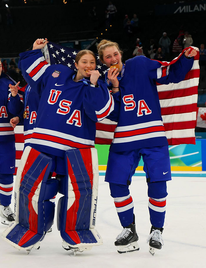 Two USA women's ice hockey players celebrating on ice, holding an American flag and biting their gold medals. Two USA women's ice hockey players celebrating on ice, holding an American flag and biting their gold medals.