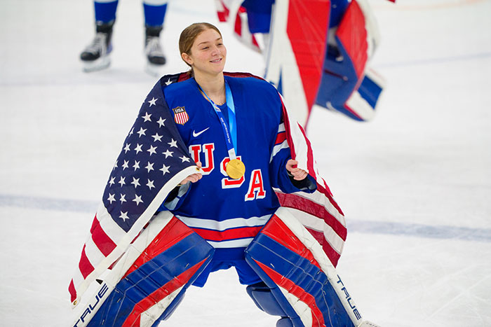 Team USA women&rsquo;s hockey star celebrating on ice with gold medal, wrapped in American flag after winning championship.