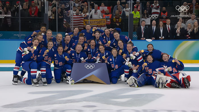 Team USA women&rsquo;s hockey stars posing on ice wearing blue uniforms after an Olympic win, celebrating with medals and smiles.