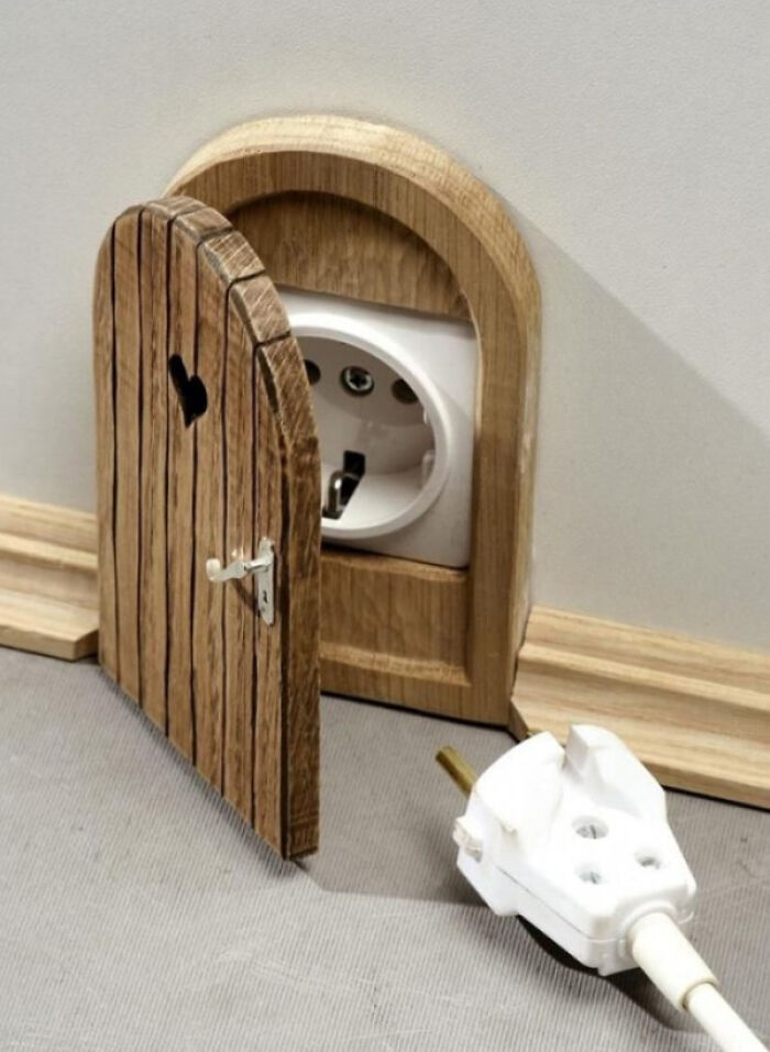 Small wooden door carved around an electrical outlet with a heart-shaped cutout and a white plug on the floor nearby.