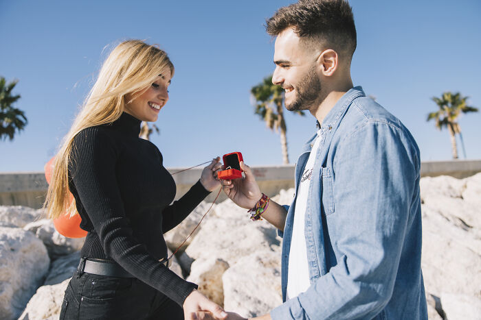 Young man proposing to a woman on the beach, both smiling amid a clear sky and palm trees, showing entitlement and surprise.