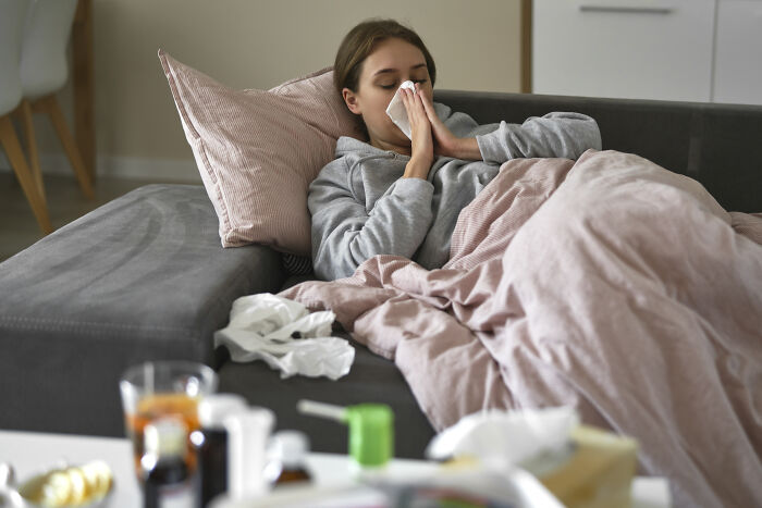 Young woman lying on couch with tissues surrounded by medicine, showing entitlement to care while feeling unwell at home.