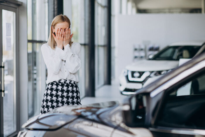 Woman covering her face in frustration standing inside a car dealership with multiple cars around, showing entitlement.