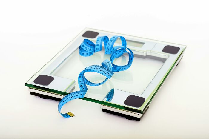 Glass digital bathroom scale with a blue measuring tape coiled on top, representing a Would You Still Do It Poll on weight.