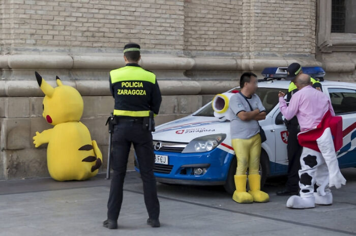 Police officer watching people in funny costumes including Pikachu and toy soldier by a police car in a humorous photo moment.