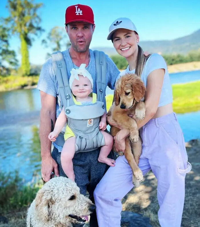 Home Improvement star Zachery Ty Bryan with family outdoors by a lake, holding a baby and two dogs on a sunny day.
