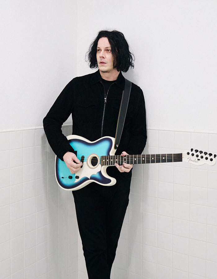 Jack White of White Stripes holding a blue electric guitar, standing against a white tiled wall in casual black clothing.