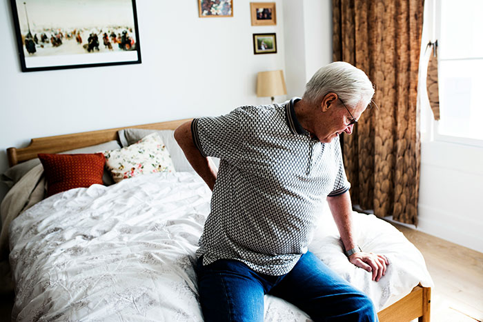 Elderly man sitting on bed holding his back in pain, highlighting husband lies about inheritance and care struggles.