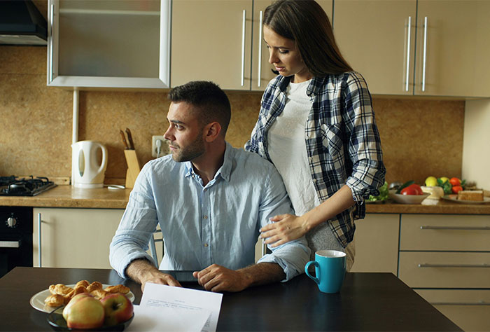 Couple in kitchen discussing finances with concern while expecting grandma to support wife as stay-at-home mom