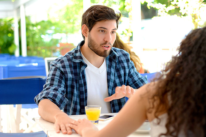 Man in plaid shirt dismissing girlfriend&rsquo;s career milestone, gesturing with hand during intense conversation at outdoor cafe.