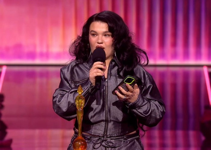 Lola Young holding an award on stage, wearing a shiny outfit resembling an oil container at a music awards event. Lola Young holding an award on stage, wearing a shiny outfit resembling an oil container at a music awards event.