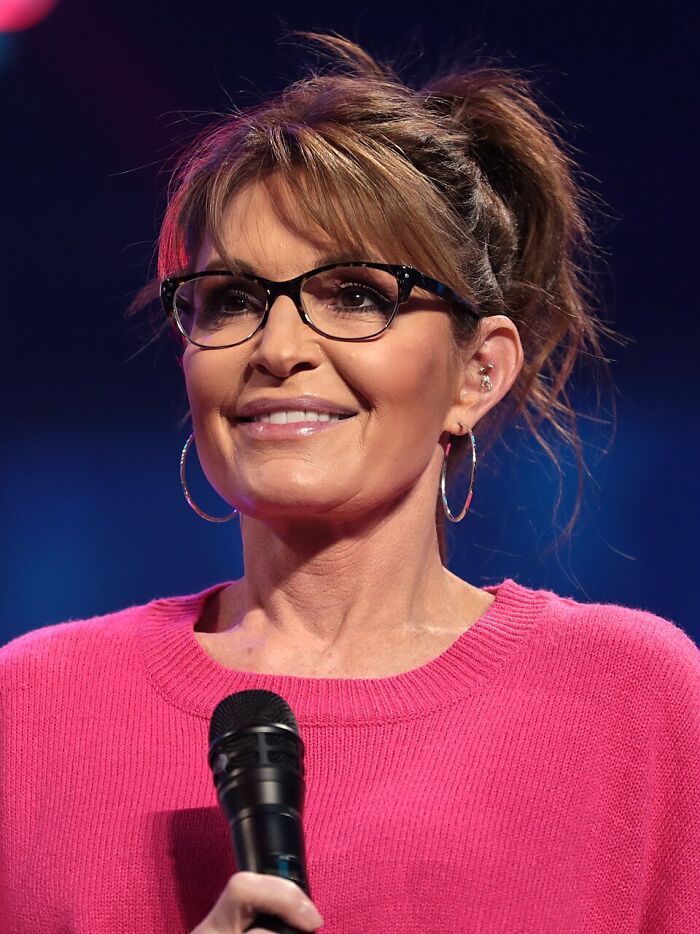 Woman wearing glasses and a pink sweater, speaking into a microphone about interesting rabbit holes in a studio setting.