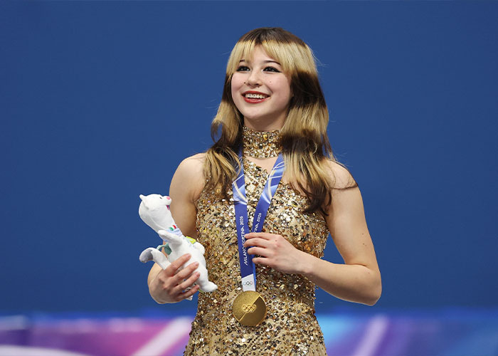 Olympic hero Alysa Liu smiling with gold medal around neck and holding a white mascot against blue background.