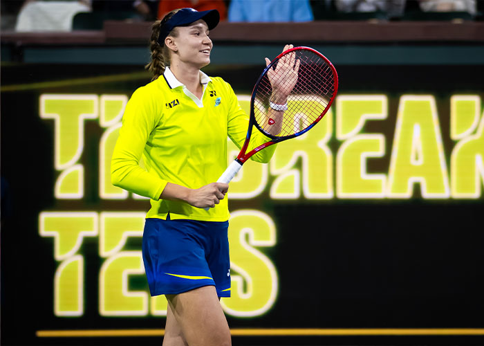 Tennis star Elena Rybakina on court wearing bright athletic wear, holding racket during trophy ceremony moment.
