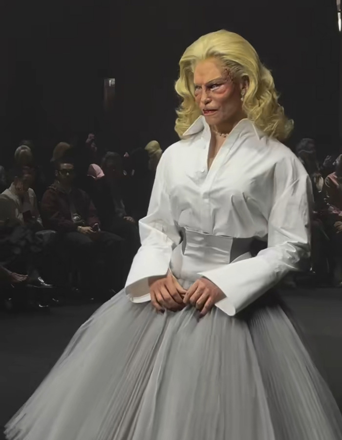 Model wearing avant-garde outfit with dramatic makeup and voluminous skirt on runway at Paris Fashion Week event.