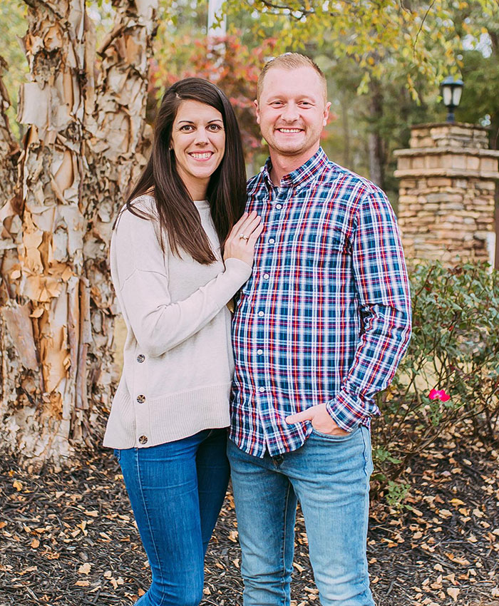 Couple standing outdoors near tree and stone pillar, representing family reaction after students prank on teacher goes tragically wrong.
