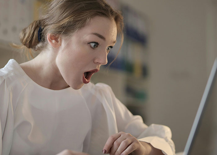 Young woman with a shocked expression looking at her laptop, reacting to messy love triangle secrets online.