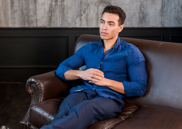 Young man in a blue shirt sitting thoughtfully on a leather couch, reflecting on relationships and men.