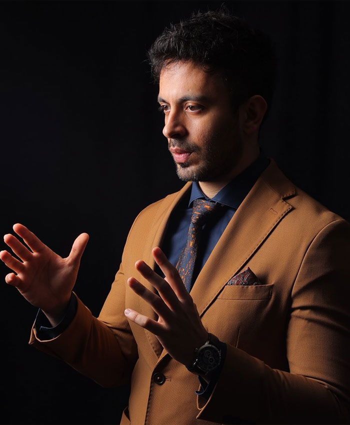Man in brown suit and patterned tie gesturing with hands against black background discussing cosmetic procedure.