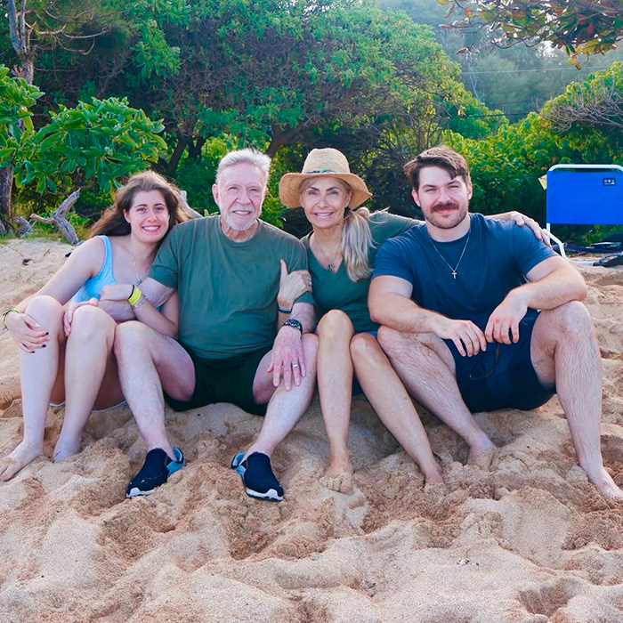 Quatro adultos sentados juntos em uma praia arenosa com vegetação ao fundo, representando os filhos de Chuck Norris quebrando o silêncio.