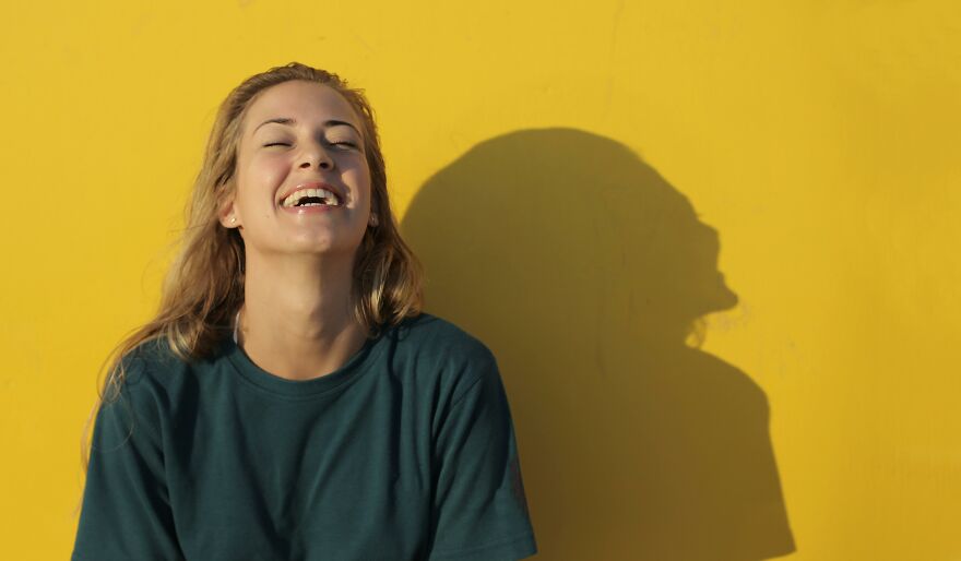 Young woman laughing happily against yellow wall showing small behaviors that make people like you more according to psychologists
