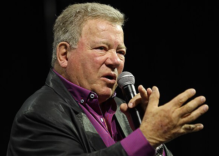 Older man speaking into a microphone during an event, illustrating stories of celebrities who turned out to be raging jerks.