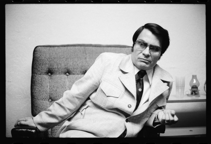 Black and white photo of a Jim Jones seated on a chair wearing glasses and a suit, reflecting historical influence.