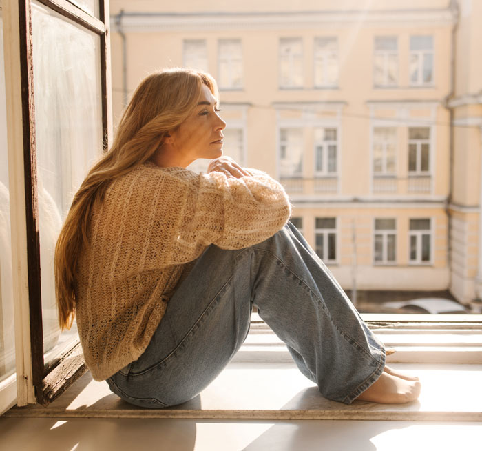 Woman sitting by an open window, lost in thought, symbolizing people who lost a friend after sharing about a tumour.