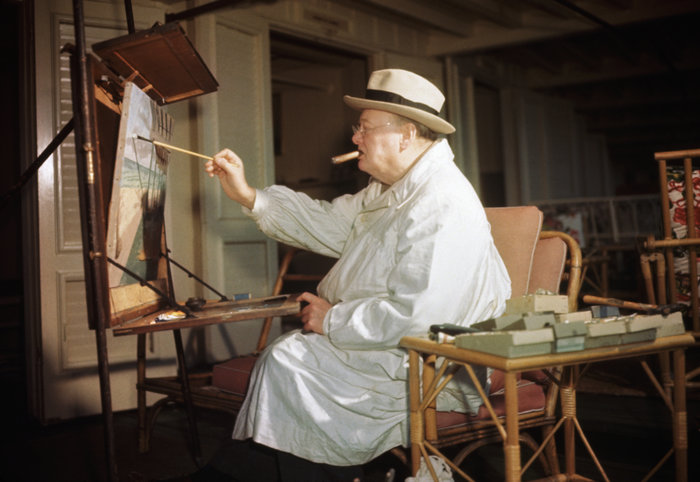 Winston Churchill painting on an easel, wearing a white coat and hat, seated in a wooden chair indoors.