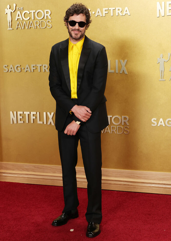 Man wearing a black suit with a bright yellow shirt and sunglasses on the red carpet at the Actor Awards event.