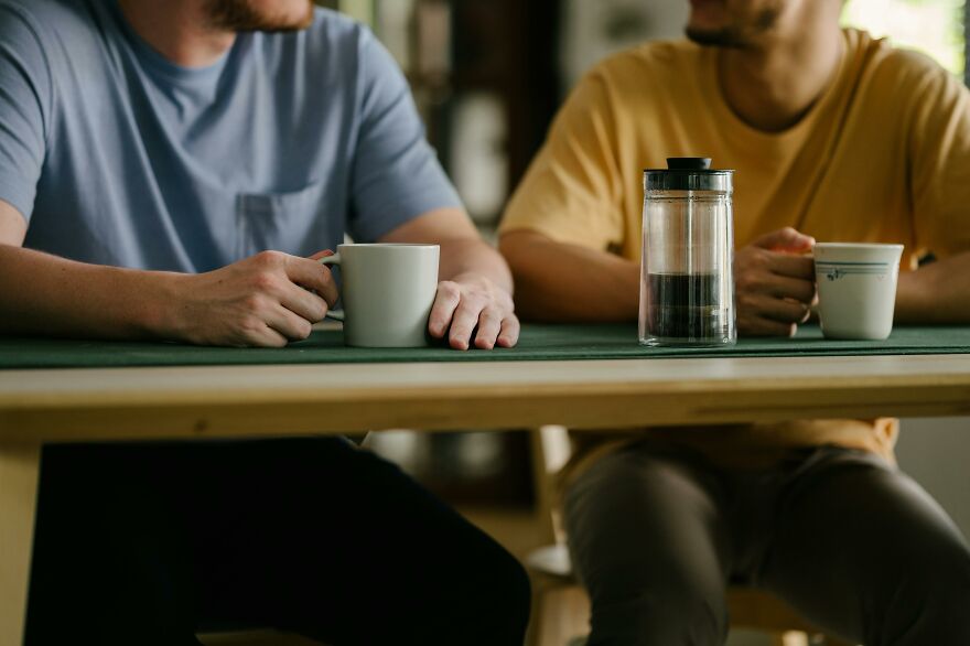 Two men sitting at a table with coffee mugs, engaging in small behaviors that make people like you more.