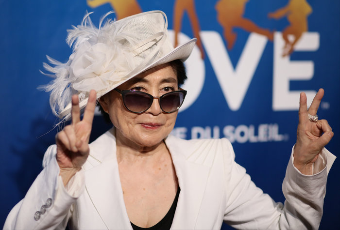 Yoko Ono wearing white suit and hat with sunglasses making peace signs at an event for famous leaders who changed history.