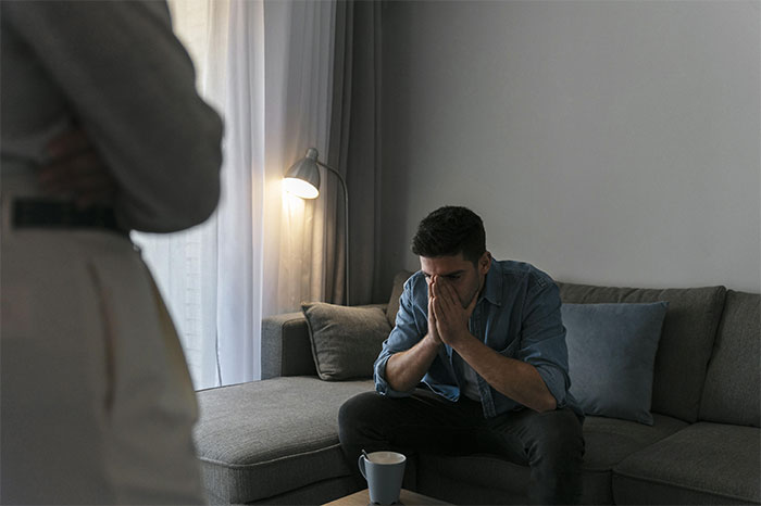 Man sitting on a couch looking distressed while another person stands nearby, depicting cheating and relationship conflict.