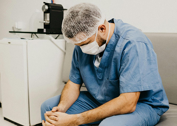 Medical worker in scrubs and mask sitting with his head down, reflecting on dark medical secrets and challenges faced.