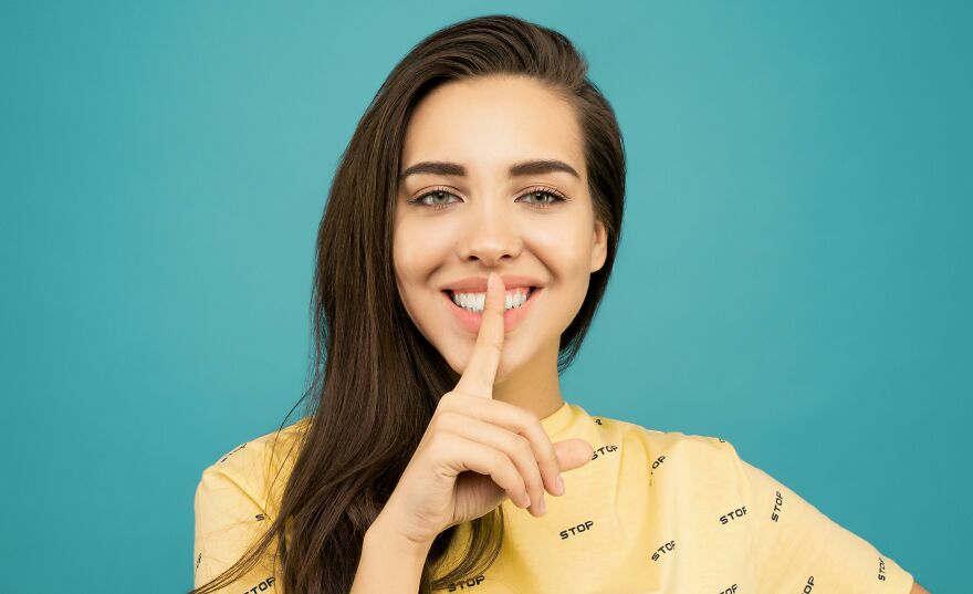 Smiling young woman in a yellow shirt making a shushing gesture showing small behaviors that make people like you more