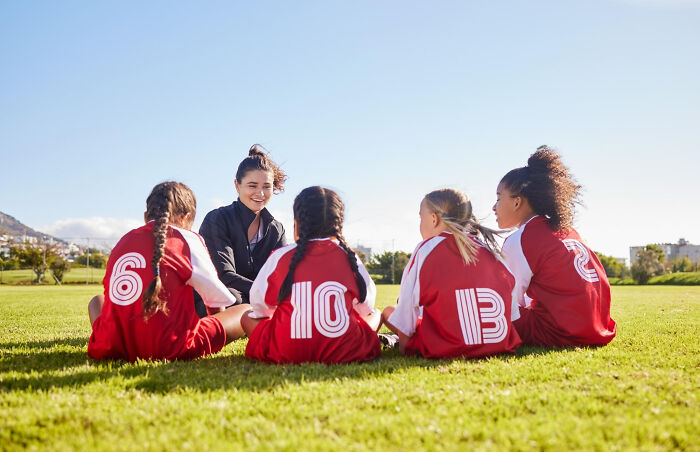 Youth soccer coach talks to young players in red jerseys sitting on grass during outdoor practice, reflecting on Gen Z stare.