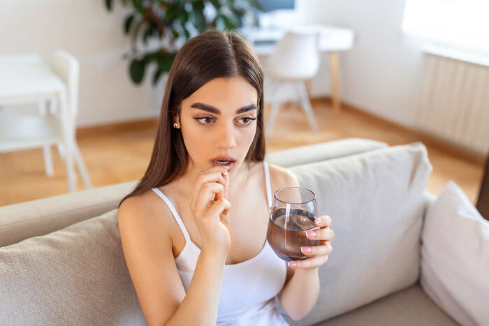 Young woman sitting on a couch taking medication with a glass of water, reflecting on women's secrets and period wardrobe.