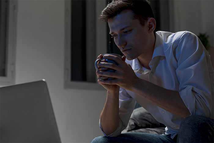 Man in a white shirt holding a mug, looking thoughtfully at a laptop in a dimly lit room about cheating and husband decisions.
